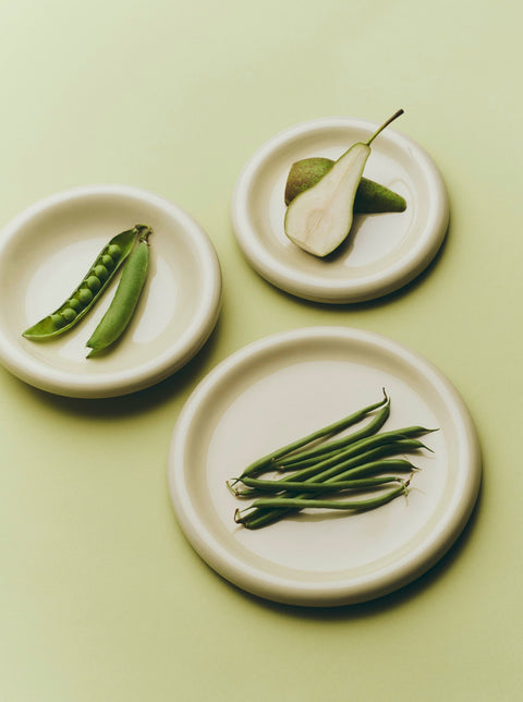 HAY Barro Bowls, Off-White, glazed terracotta bowls and plates, overhead with pea pods, a halved pear and green beans on pale green