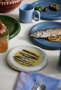 HAY Barro Cups, Light Blue, terracotta cups, one cup on a laid table with sardines, a grilled fish on a blue plate and a lavender napkin