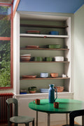 HAY Barro Cups, Natural, terracotta cups, one cup on a green round table in a kitchen with shelves of stacked plates and a blue jug