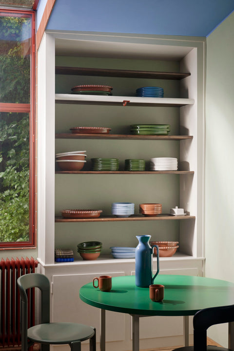 HAY Barro Cups, Natural, terracotta cups, one cup on a green round table in a kitchen with shelves of stacked plates and a blue jug