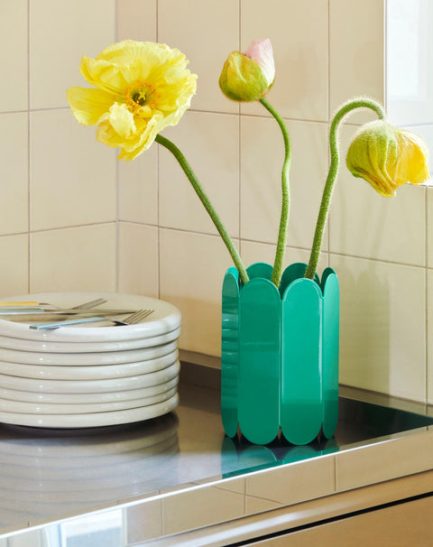 HAY Arcs Vase, Small, Green, green metal flower vase with scalloped sides, holding yellow poppies on a steel counter beside a stack of plates