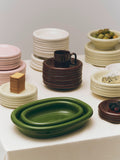 HAY Barro Oval Dish, Small, Green, glazed ceramic oval serving dish, two green dishes with stacks of plates and bowls on a pale table