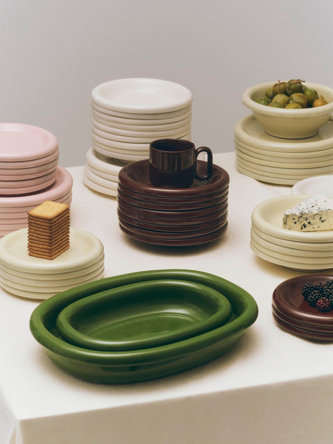 HAY Barro Oval Dish, Small, Green, glazed ceramic oval serving dish, two green dishes with stacks of plates and bowls on a pale table