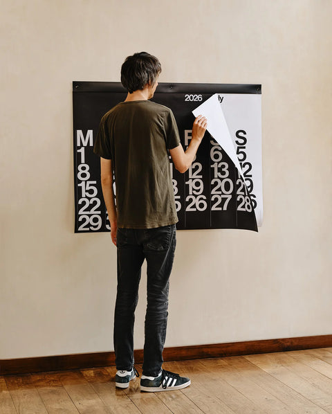 Person flipping a page on the 2026 Stendig Calendar mounted on a wall