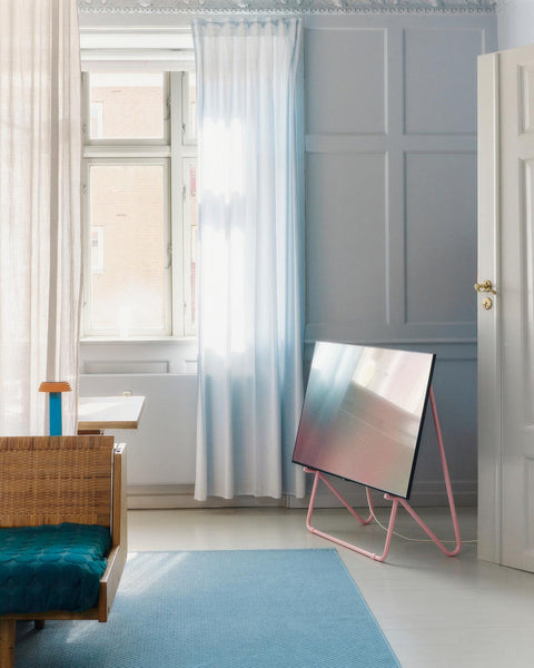 Pedestal Bloom TV Stand in Bubble Gum, VESA-free leaning TV stand, holding a screen in a bedroom with a blue rug and a window behind