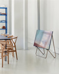 Pedestal Bloom TV Stand in Sandstorm, VESA-free leaning TV stand, holding a screen in a white room beside a dining table and wooden chairs