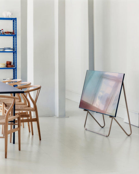 Pedestal Bloom TV Stand in Sandstorm, VESA-free leaning TV stand, holding a screen in a white room beside a dining table and wooden chairs