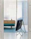 Pedestal Bloom TV Stand in Chrome, VESA-free leaning TV stand, seen from behind through a doorway into a blue room with a bench