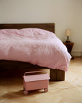 Pedestal Cable Villa in Bubble Gum, steel cable box on wheels, standing on a wooden floor beside a bed