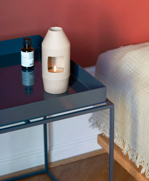 HAY Chim Chim Scent Diffuser, Light Beige, ceramic tealight scent diffuser, lit on a blue tray table beside a bed and a red wall