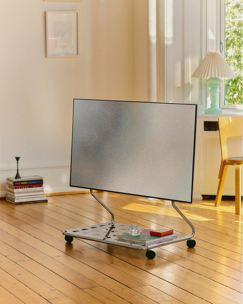 Pedestal Disco TV Stand in Chrome, low stand on wheels holding a flat screen in a bright room with a wooden floor
