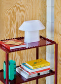 HAY Arcs Vase, Small, Green, green metal flower vase with scalloped sides, on the middle shelf of a deep red trolley with books and a white lamp
