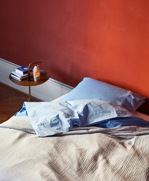 HAY Chim Chim Scent Diffuser, Light Beige, ceramic tealight scent diffuser, lit on a side table by a bed with blue linen and a red wall