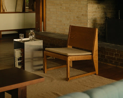 Frama Easy Chair 01 in Warm Brown Birch, birch low chair with a pale seat cushion beside a brick fireplace and a metal side table