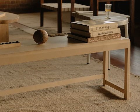 Frama Bench 01 in Natural Birch, pale birch bench on a rug carrying a stack of books and a wooden ball, table legs behind