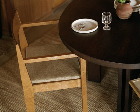 Frama Armrest Chair 01 Cushion in Camel, woven seat pad on a birch armchair at a dark round table set with a plate and a glass