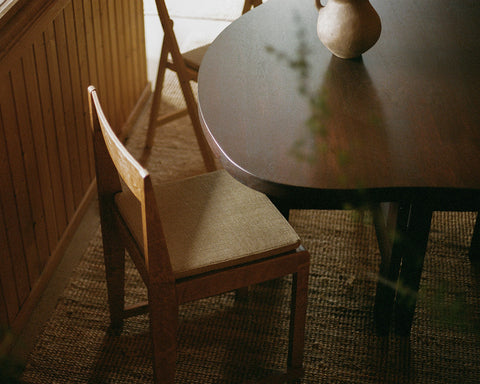 Frama Chair 01 Cushion in Camel, woven seat pad on a wooden chair pulled up to a dark round table on a woven rug
