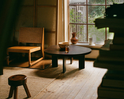 Frama Farmhouse Coffee Table in Dark Oak, dark table by a window holding a bowl and a vase, wooden chair and stool on a sisal rug