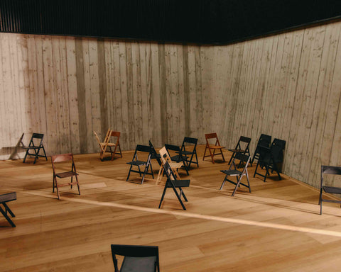 Frama Folding Flat Chair in Ash Black Birch, rows of black, brown and pale folding chairs set out across a plywood lined hall