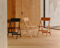 Frama Folding Flat Chair in Natural Birch, pale folding chair between a black one and a warm brown one against a pine clad wall