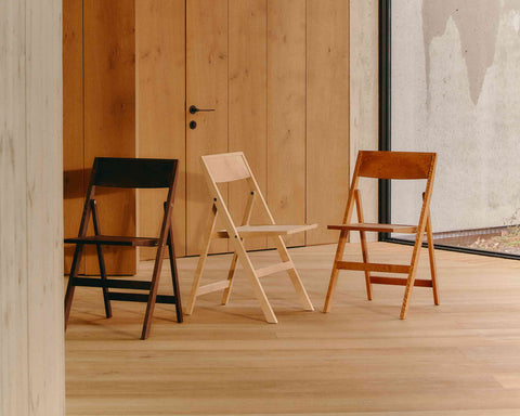 Frama Folding Flat Chair in Natural Birch, pale folding chair between a black one and a warm brown one against a pine clad wall