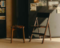 Frama Folding Flat Chair in Ash Black Birch, black folding chair open before a dark cabinet pinned with photos, stool alongside