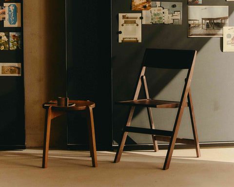 Frama Folding Flat Chair in Ash Black Birch, black folding chair open before a dark cabinet pinned with photos, stool alongside