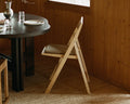 Frama Folding Flat Chair Cushion in Camel, woven pad on a pale folding chair beside a dark round table in a pine panelled room