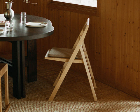 Frama Folding Flat Chair Cushion in Camel, woven pad on a pale folding chair beside a dark round table in a pine panelled room