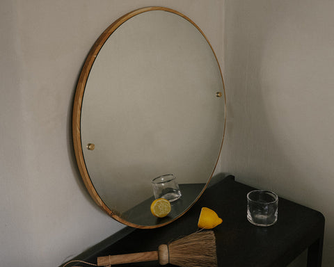 Frama CM-1 Circle Mirror, oak rimmed mirror on a grey wall above a black table holding a glass, a lemon and a brush