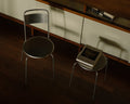 Frama Petit Rond Stool in Stainless Steel, stool with a book on its seat next to a matching steel chair on a dark wood floor