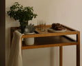 Frama Ratio Trolley in Warm Brown Birch, birch trolley in a room corner holding a vase of greens, a glass and a tray of tomatoes