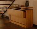 Frama Ratio Chest in Warm Brown Birch, chest with the top drawer pulled open, set under a staircase with books and a dark jug on top