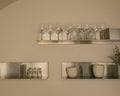 Frama Rivet Shelf, three metal shelves on a kitchen wall, one holding a row of wine glasses with bowls and tumblers below