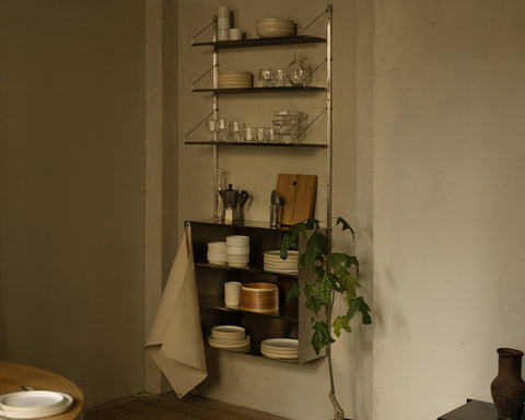 Frama Shelf Library Canvas Cabinet Section in Stainless Steel, dim kitchen corner with plates, glassware and a moka pot on the steel shelves