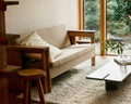 Frama Symmetry Couch in Oat Fabric and Ash, couch on a shaggy rug by a garden window with a cushion, a wooden stool and a low dark table