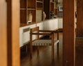 Frama Symmetry Easy Chair in Oat Fabric and Honey Ash, chair on a dark wood floor seen through a doorway, wooden shelving behind it