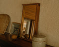 Frama Symmetry Mirror, mirror standing on a dark shelf against a rough yellow wall beside a woven fan and a speckled ceramic jar