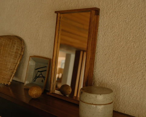 Frama Symmetry Mirror, mirror standing on a dark shelf against a rough yellow wall beside a woven fan and a speckled ceramic jar
