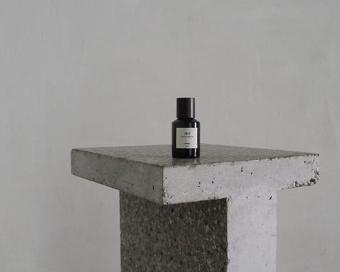Frama T-Table in Terrazzo, table against a grey wall with a small dark glass bottle standing on the speckled slab top