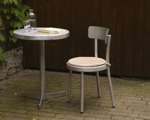 Frama Tasca Cushion in Beige, pad tied to an aluminum chair seat on brick paving beside a round table with a bottle and a book