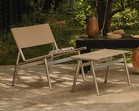 Frama Union Stool in Sandstone Linen and Aluminum, stool and matching easy chair on a paved terrace with a bench and trees behind