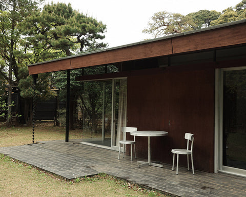 Frama Tasca Chair in Aluminum, two chairs and a small round table on the timber deck of a dark wood house among pine trees
