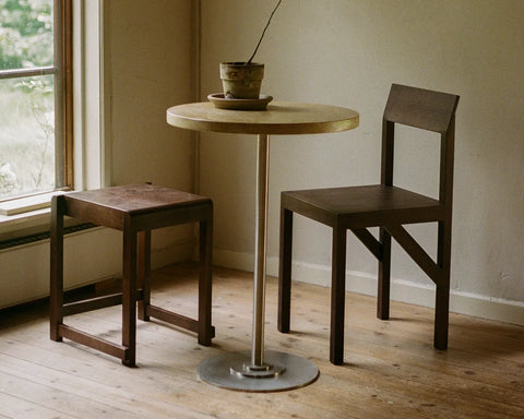 Frama Low Stool 01 in Dark Brown Birch, dark stool beside a round pedestal table and a chair in a room with a plank floor