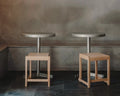 Frama Low Stool 01 in Natural Birch, two pale birch stools tucked under round metal cafe tables against a mottled grey wall