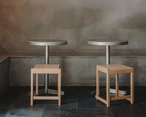 Frama Low Stool 01 in Natural Birch, two pale birch stools tucked under round metal cafe tables against a mottled grey wall