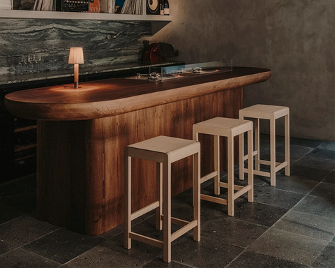 Frama Stool 01 in Natural Birch, three pale birch stools lined up at a dark wood kitchen island with a small lamp on top