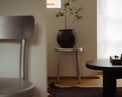 Frama Tasca Stool in Aluminum, stool holding a dark ceramic vase of branches in a corner beside a blurred chair and dark table