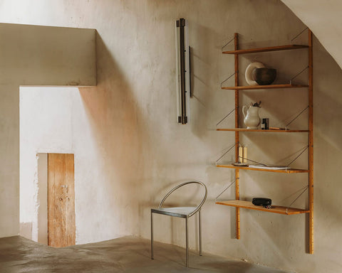 Frama Triangolo Chair in Stainless Steel, chair standing in a plaster walled room beside an oak wall shelf holding jugs and ceramics