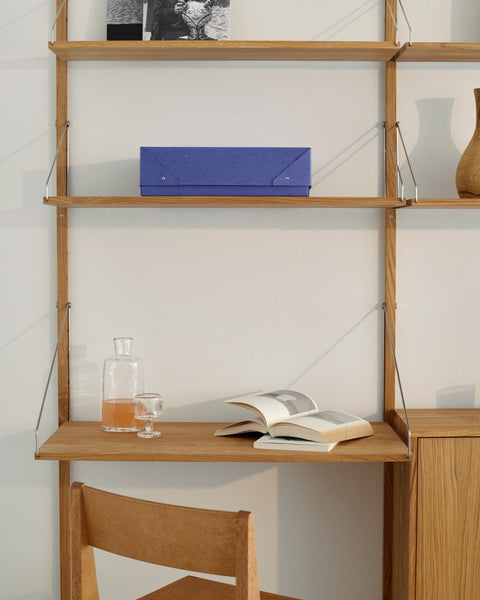 Frama Shelf Library in natural oak mounted in a corner nook with ceramics, a table lamp, and an aluminum Rivet cart, beside a Frama Chair 01